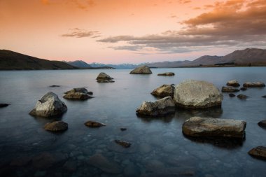 Gün batımında sakin bir göl manzarası sakin sular ve dağılmış kayalar yumuşak, pastel bir gökyüzü altında uzak dağlar tarafından çerçevelenmiş..