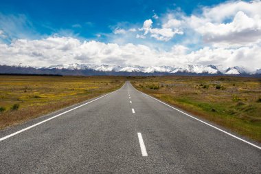 Uzun, düz bir yol uzanan karlı dağlara doğru canlı mavi bir gökyüzünün altında, açık arazinin geniş güzelliğini yakalıyor..
