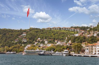 Bir yaz günü, rıhtımdaki tekneler, geleneksel evler, sık ağaçlar ve büyük Türk bayrağıyla, Avrupa 'nın Boğaz tarafındaki yeşil dağların manzarasına bakın.