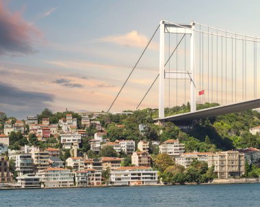 İstanbul, Türkiye 'deki İstanbul Boğazı' nın Avrupa yakası denizinden İstanbul 'a, İstanbul köprüsünün altında bulunan geleneksel rıhtım evleri ve yoğun ağaçlı yeşil tepeye güneş doğduktan sonraki yaz günü bakın.