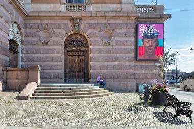 Stockholm, İsveç - 26 Haziran 2022: Tuğla taş duvarın önündeki merdivenlerde kitap okuyan yaşlı bir adam, reklam panosu 3. Hikayeler ve Kraliyet Opera Binası 'na açılan ahşap kapı
