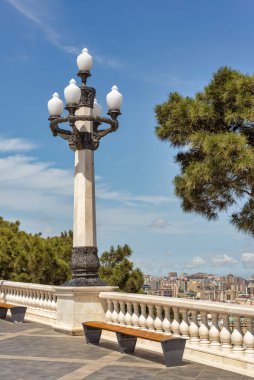 Süslü siyah metal detayları olan beyaz taştan bir sokak direği, uzaktaki ağaçların ve binaların olduğu bir şehre bakan taş bir geçitte duruyor.