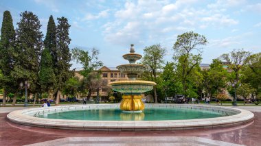 Baku, Azerbaijan - May 5, 2024: In Bakus Sahil Bagi park, a golden based fountain with blue and white tiles sits in a circular pool, surrounded by lush trees and a walkway clipart