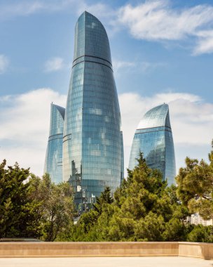 Bakü, Azerbaycan 'daki İkonik Ateş Kuleleri' nin Martyrs Lane manzarası alevlere benzeyen mavi gökyüzüne karşı dik duruyor. Ön plandaki ağaçlar şehir manzarasına doğal bir dokunuş ekler.