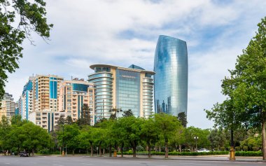Bakü, Azerbaycan 'daki modern yüksek binaların manzarası, JW Marriott Absheron oteli de dahil olmak üzere ön planda bir sıra ağaç ve açık mavi gökyüzü.