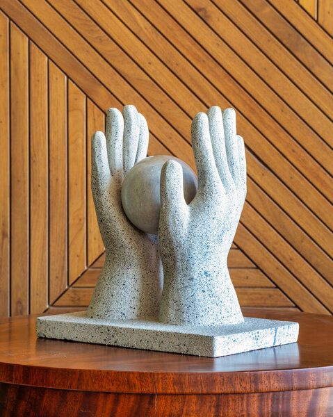 Каменная скульптура изображает две руки, мягко колышущиеся на круглом шаре, установленном на деревянном столе со стильным геометрическим деревянным фоном