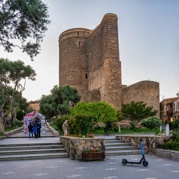 stock image Baku, Azerbaijan - May 9 2024: The Maidan Tower in the Old City stands tall against a backdrop of clouds in the evening sky. Lush greenery surrounds the historic structure.