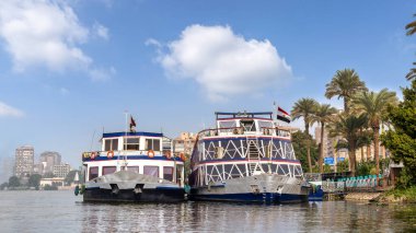 Kahire, Mısır - 9 Aralık 2023: Nile Crystal Cruise gemisi Maadi bölgesinde Nile Corniche boyunca yanaştı. Bereketli palmiye ağaçları ve kentsel mimari açık mavi gökyüzünün altında canlı bir zemin oluşturur.