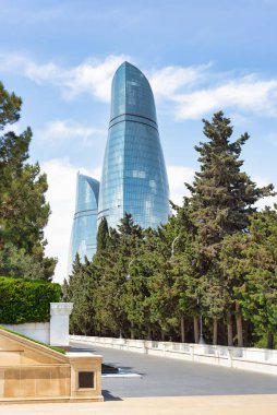 Martyrs Lane 'den, Bakü, Azerbaycan' daki ikonik Alev Kuleleri açık mavi gökyüzüne karşı görkemli bir şekilde yükselirken, şekilleri alevleri andırırken, ön plandaki ağaçlar kentsel manzarayı güçlendiriyor.