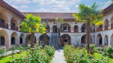 Shaki, Azerbaycan 'daki Yukarı Caravanserai' nin avlusunda, ziyaretçileri bu dingin manzaranın tadını çıkarmaya davet eden kavisli balkonları, palmiye ağaçları ve canlı çiçekleri olan çarpıcı bir mimari bulunuyor.