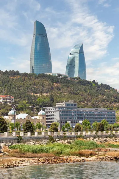 İkonik Alev Kuleleri, Azerbaycan 'ın Bakü siluetine hakim. Ağaçlarla çevrili ve bir liman bölgesi olan manzaranın üzerinde görkemli bir şekilde yükselen güneş ışığını yansıtıyor.