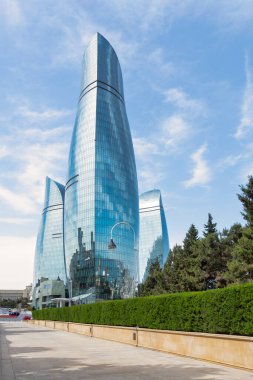 Bakü, Azerbaycan 'daki ikonik Alev Kuleleri Martyrs Lane' den görüldüğü üzere mavi gökyüzüne karşı görkemli bir şekilde yükseliyor. Ön plandaki ağaçlar doğal bir element ile şehir manzarasını güçlendirir.