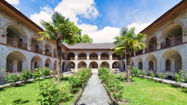 Shaki, Azerbaycan 'daki Yukarı Caravanserai avlusu, muhteşem bir mimariye sahip kemerli balkonlar, palmiye ağaçları ve canlı çiçeklerle, dingin bir ortam yaratıyor.