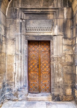 Güzel tasarlanmış ahşap bir kapı, yıpranmış taştan bir duvarın içine yerleştirilmiş, tarihi mimari ve işçiliğin sergilendiği detaylı geometrik desenlere sahiptir.
