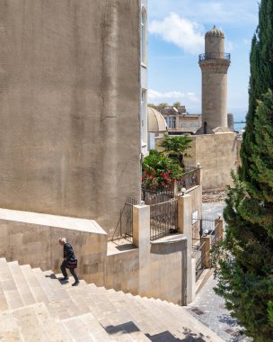 Bakü, Azerbaycan - 10 Mayıs 2024: Yaşlı adam tarihi Beyler Camii 'nin minaresi ve arkadaki büyüleyici caddelerle Eski Şehir Icherisheher' de merdivenlerden çıktı