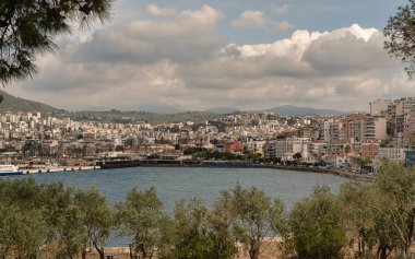 Türkiye 'de Aydın' ın Kusadasi ilçesinin deniz manzarası ve şehir manzarası. Yeşil ağaçlar ön planda yer alırken, canlı binalar kıyı boyunca uzanıyor.