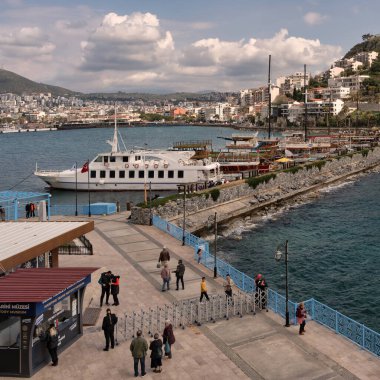 Aydın, Türkiye - 3 Nisan 2025: Kusadasi Şatosu 'na bakan Kusadasi Limanı, parçalı bulutlu gökyüzü altında kıyı güzelliğinin keyfini çıkaran tekne ve ziyaretçilerle dolup taşıyor