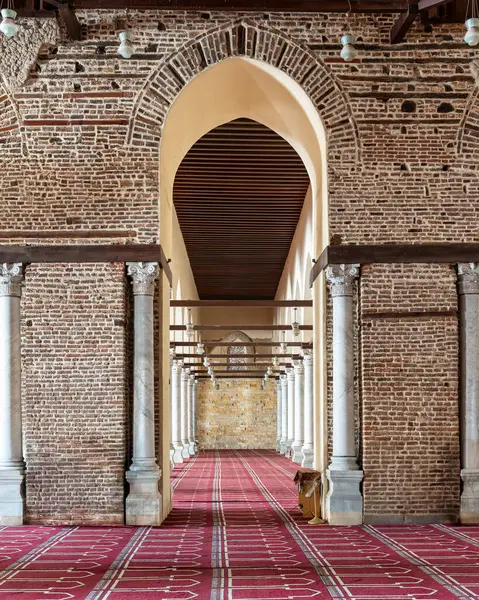 Mısır 'ın Kahire kentindeki tarihi Al-Zahir Baybarları Camii' nin iç manzarası, bir kemer yoluyla, tuğla duvarlar, mermer sütunlar ve koridorun aşağısına uzanan uzun kırmızı bir dua halısı.