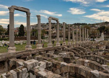 İzmir, Türkiye - 1 Nisan 2025: İzmir Agorası. Bu tarihi arkeoloji sahasında, arkasında dağlar ve şehir binaları olan yeniden inşa edilmiş Roma sütunları ve kadim kemerli temeller.