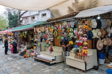 Selcuk, Türkiye - 3 Nisan 2025: Sirince Köyü 'ndeki hareketli sokak pazarı, renkli örülü anahtarlıklar ve hasır şapkalarla dolu bir tezgahta bir satıcı yer alıyor