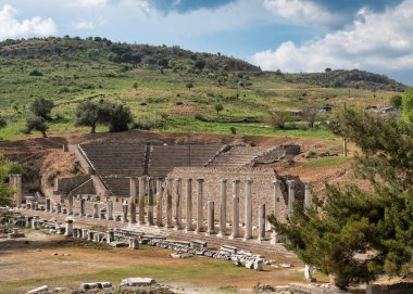 Türkiye 'nin Bergama kentindeki Antik Asklepion kalıntıları. Roma tiyatrosu ve kutsal yol sütunları bulutlu bir gökyüzünün altındaki yeşil tepelerin arasında. Tarihi dönüm noktası