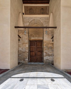 Kahire, Mısır 'daki al-Zahir Baybarları Camii' nin süslü girişinin görüntüsü. Eski ahşap kapılar, taş kemer yolu, karmaşık oymalar ve dramatik gölgeli mermer zemin. İslami Miras