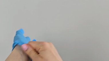 Mavi tıbbi eldiven giyip avuçlarını gösteriyor. Eller dikkatli bir şekilde tek kullanımlık nitril eldivenler giyer ve sonra da eldivenlerdeki avuç içlerini gösterir. Arka plandaki gri duvar, temiz ve profesyonel tıbbi ortamı güçlendiriyor.. 