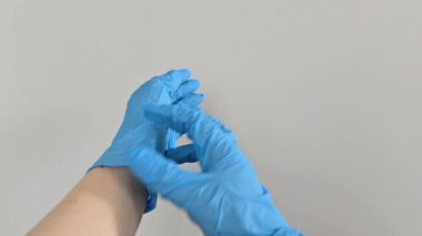 Bilekten mavi tıp eldivenlerini çıkarıyorum. Eller, hijyen ve güvenliğe odaklanarak tek kullanımlık nitril eldivenleri çıkarır. Arka plandaki gri duvar tıbbi çevreyi vurguluyor.