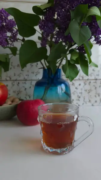 Mor leylak dalları açan vazo, bir fincan fındık, iki elma ve mutfak masasında bir bardak sıcak çay. Arka planda tutacağı olan bir mutfak duvarı. Sıcak bahar durgun hayat.