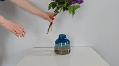 Mor eflatun dalları, açık gri duvarlı beyaz bir masanın üzerindeki mavi ve sarı cam bir vazoya yerleştirmek. Yatay görüntüde minimalist çiçek düzeneği.