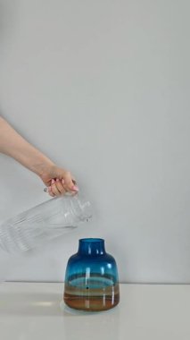 Beyaz masadaki açık gri duvarlı mavi ve sarı cam vazo. El cam sürahiden cama su döküyor, masaya da biraz su dökülüyor. Minimalist dikey video sahnesi.
