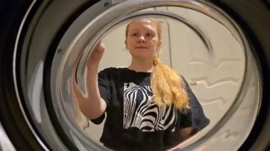 Çamaşır makinesinin içinden bir kadının kapıyı açtığını ve çamaşırları yüklediğini gösteren bir görüntü. Yakın çekim, günlük ev işlerini yakalıyor ve rahat bir evde bakım görüyor..