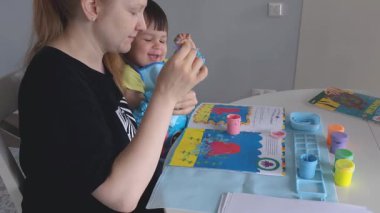 Anne ve bebek evde parmak boyalarıyla resim yapıyorlar. Yaratıcı kaynaşma zamanı, koruyucu önlüklü ve kollu çocuk. Eğlenceli dağınık oyun ve erken dönem sanat gelişimi aktivitesi.