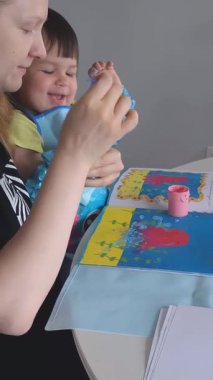 Anne ve bebek evde parmak boyalarıyla resim yapıyorlar. Yaratıcı kaynaşma zamanı, koruyucu önlüklü ve kollu çocuk. Eğlenceli dağınık oyun ve erken dönem sanat gelişimi aktivitesi.