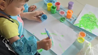 Anne ve bebek evde parmak boyalarıyla resim yapıyorlar. Yaratıcı kaynaşma zamanı, koruyucu önlüklü ve kollu çocuk. Eğlenceli dağınık oyun ve erken dönem sanat gelişimi aktivitesi.