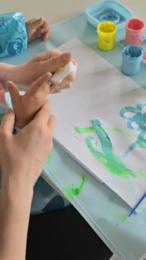 Anne ve bebek evde parmak boyalarıyla resim yapıyorlar. Yaratıcı kaynaşma zamanı, koruyucu önlüklü ve kollu çocuk. Eğlenceli dağınık oyun ve erken dönem sanat gelişimi aktivitesi.