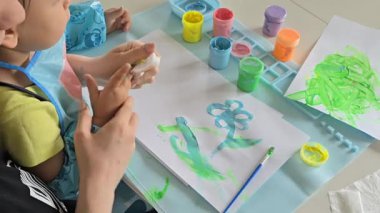 Anne ve bebek evde parmak boyalarıyla resim yapıyorlar. Yaratıcı kaynaşma zamanı, koruyucu önlüklü ve kollu çocuk. Eğlenceli dağınık oyun ve erken dönem sanat gelişimi aktivitesi.