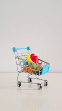 Küçük alışveriş arabası beyaz bir masada küçük oyuncak poşetleriyle dolu. Alışveriş, tüketici kültürü ve günlük aile yaşantımızın minyatür bir temsili..