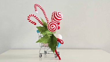 Noel süsleri, kırmızı şeker kamışları, lolipoplar ve ökse otu yapraklarıyla dolu mini alışveriş arabası. Neşeli bir tatil alışverişinin ve mevsimlik ruhun sembolik bir görüntüsü..