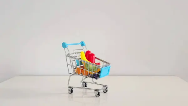 Küçük alışveriş arabası beyaz bir masada küçük oyuncak poşetleriyle dolu. Alışveriş, tüketici kültürü ve günlük aile yaşantımızın minyatür bir temsili..