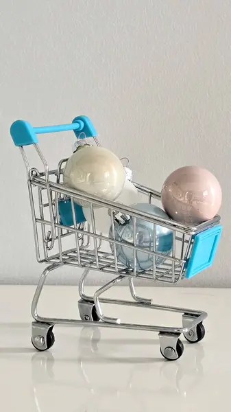 Pastel Noel süsleriyle dolu mini alışveriş arabası beyaz masa ve gri arka plan tatil alışverişini, festival hazırlıklarını ve mevsimlik dekorasyonu simgeliyor..