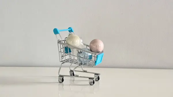 Pastel Noel süsleriyle dolu mini alışveriş arabası beyaz masa ve gri arka plan tatil alışverişini, festival hazırlıklarını ve mevsimlik dekorasyonu simgeliyor..