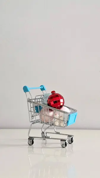 Pastel Noel süsleriyle dolu mini alışveriş arabası ve üstünde Noel alışverişinin, sezonluk dekorun ve renk ve ruh halindeki kontrastın bayram sembolü olan kırmızı bir mücevher..