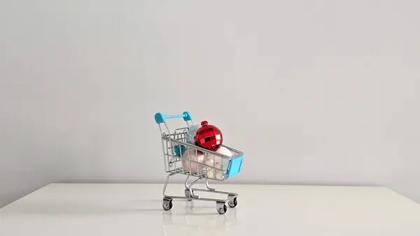 Pastel Noel süsleriyle dolu mini alışveriş arabası ve üstünde Noel alışverişinin, sezonluk dekorun ve renk ve ruh halindeki kontrastın bayram sembolü olan kırmızı bir mücevher..