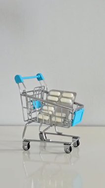 Dikey yakın çekim videosu: bir el, günlük sağlık hizmetleri ve kişisel sağlığı sembolize eden yumuşak gri arka planlı beyaz bir zemindeki küçük bir alışveriş arabasının içinde su toplayan hapları yerleştiriyor.