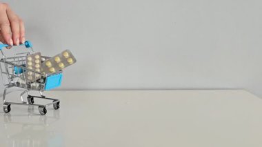 Su toplayan haplarla dolu mini bir alışveriş arabası beyaz bir masanın üzerindeki çerçeveye itiliyor. Gri arka planı sağlık, ilaç ve günlük sağlık hizmetlerini temsil ediyor..