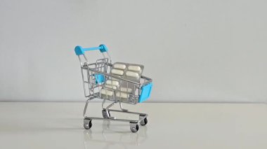 Bir el, ilaç paketlerini küçük bir alışveriş arabasına yerleştiriyor. Gri bir geçmişi olan beyaz bir masaya. Tıbbi harcamaları, sağlık giderlerini sembolize ediyor..