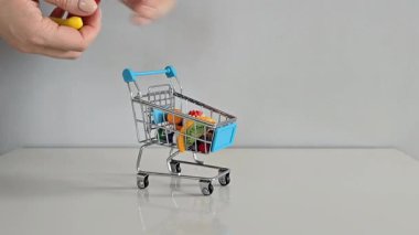 Bir el, aile gıda alışverişini, yükselen fiyatları ve günlük giderleri simgeleyen gri arka planı olan beyaz bir masanın üzerindeki mini alışveriş arabasından oyuncak poşetleri alıyor..