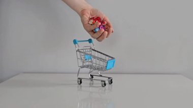 Bir el, renkli değerli taşlar ve Noel ıvır zıvırı şeklindeki şenlikli lastik eşyalarla dolu küçük bir oyuncak arabasını gri bir arka planı olan beyaz bir masanın üzerine koyar..