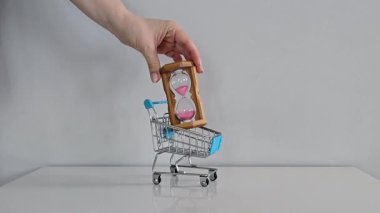 Gri arka planı olan beyaz bir masada mini oyuncak alışveriş arabası. Bir el, içine zamanı, aciliyeti ve tatil geri sayımını simgeleyen bir kum saati koyar..